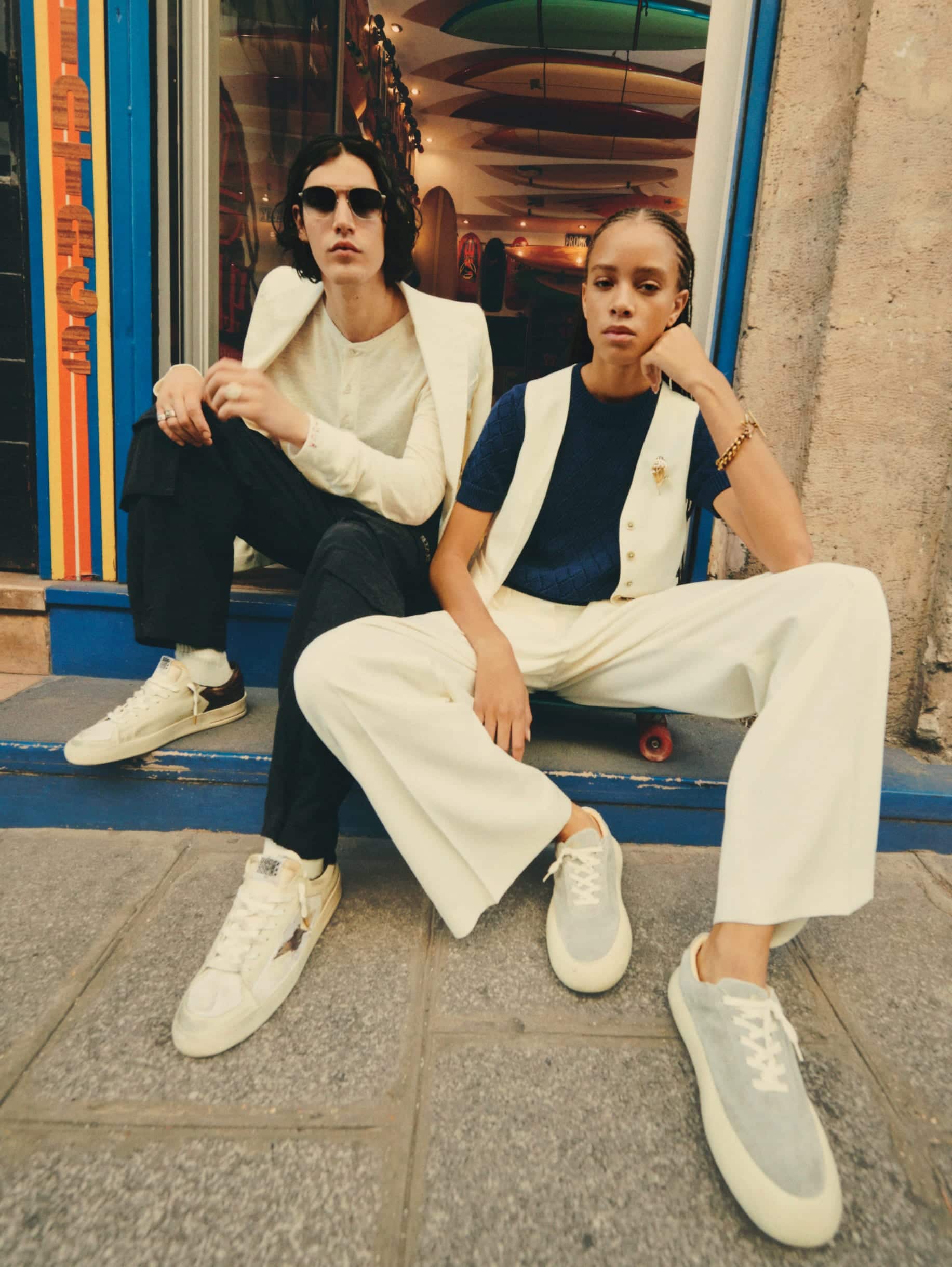 Man-with-glasses-and-stardan-sneakers-sitting-on-porch-of-skateboard-shop-with-woman-wearing-white-pants-and-ice-grey-space-star-sneakers