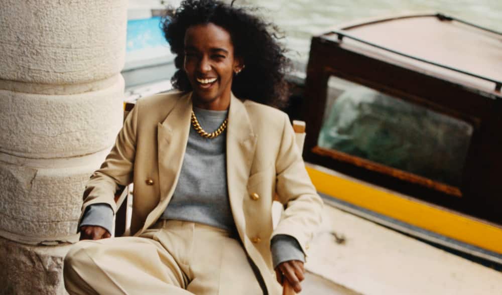 woman-wearing-beige-jacket-and-pants-sitting-next-to-white-column-with-boat-behind
